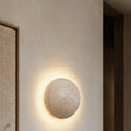 Kasumi wall light, a natural travertine disc casting a soft halo on the wall