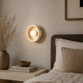 Wa wall light, a round natural alabaster lamp glowing beside a bed